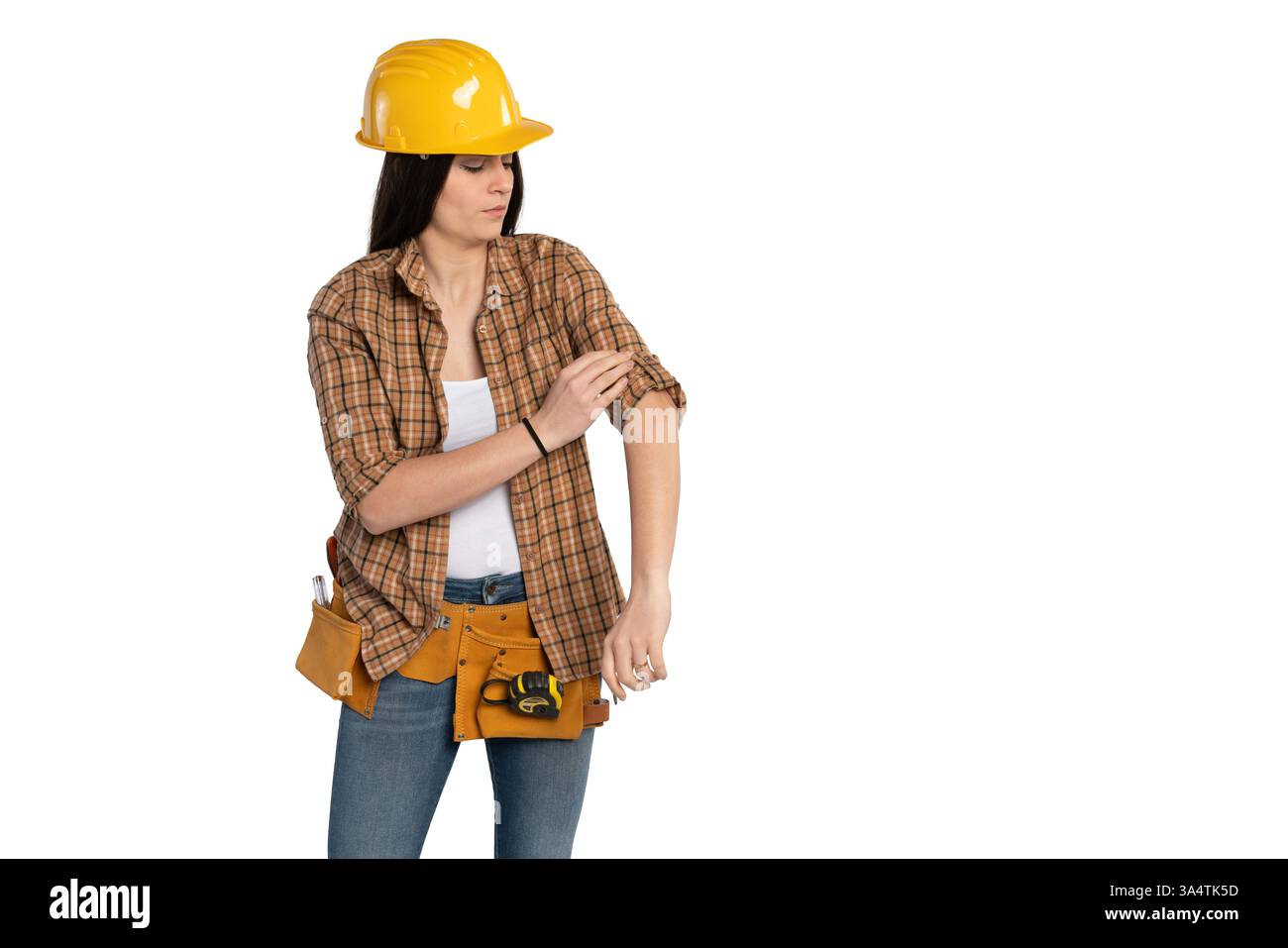 Giovane lavoratrice edile con un elmetto giallo e una camicia a quadri che si arrotola sulle maniche, preparandosi al lavoro. Indossa una cintura portautensili con attrezzi, je Foto Stock