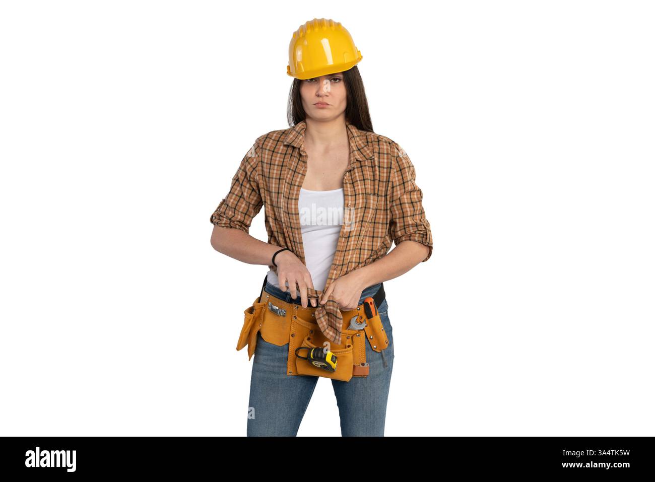 Una lavoratrice edile con un elmetto giallo e una camicia a quadri che regolano la cintura degli attrezzi. Indossa jeans e una canotta bianca, in piedi con fiducia. Foto Stock