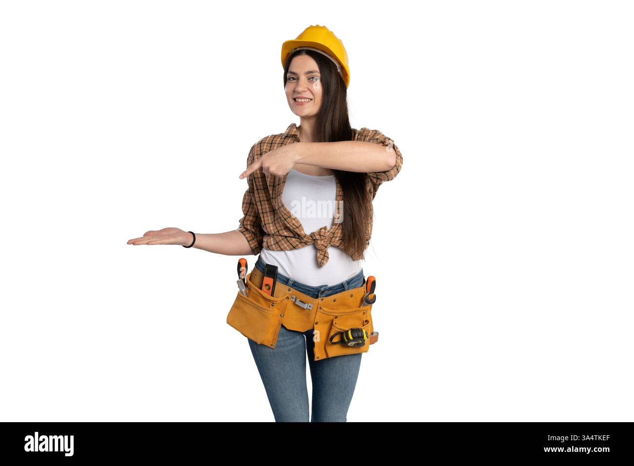 Una lavoratrice edile sorridente che indossa un elmetto giallo e una cintura portautensili punta sul palmo aperto, come se presentasse un prodotto o un'idea. Lei è una Foto Stock