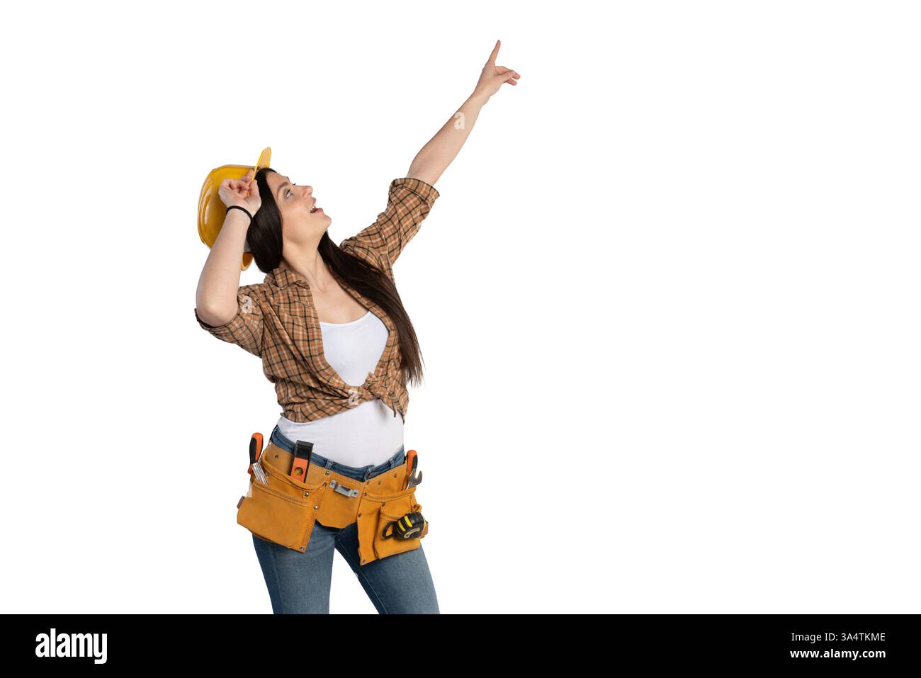 Una lavoratrice edile sicura con una camicia a quadri e una cintura portautensili punta verso l'alto tenendo il suo elmetto giallo. Isolato su sfondo bianco Foto Stock