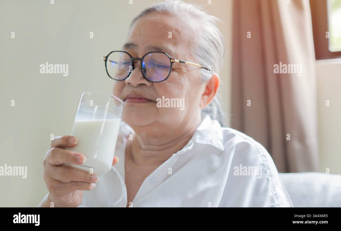 Felice sorridente donna asiatica anziana che beve latte caldo o integratori alimentari dal bicchiere. Messa a fuoco selettiva. Foto Stock