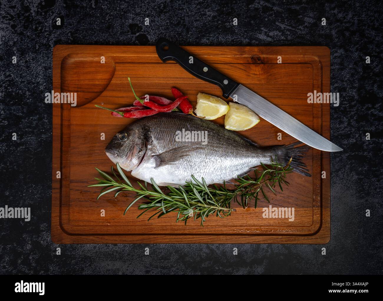 Orata con testa dorata su tagliere in legno (dorata), limone e rosmarino su sfondo scuro Foto Stock