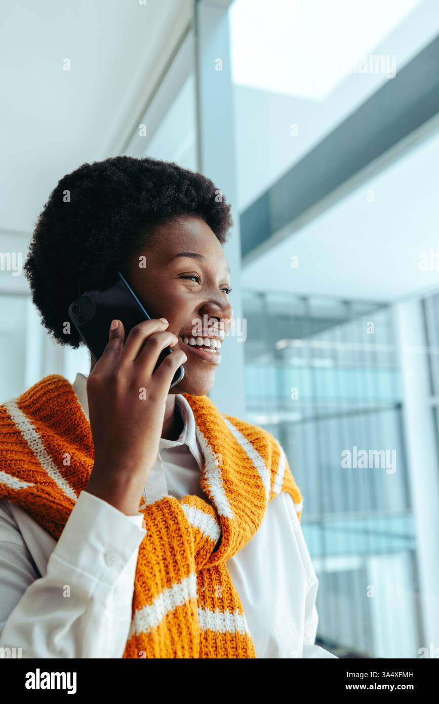 Una donna africana professionista sorridente, con un elegante top arancione, è impegnata in una telefonata all'interno di un ufficio contemporaneo. Incarna fiducia e.. Foto Stock