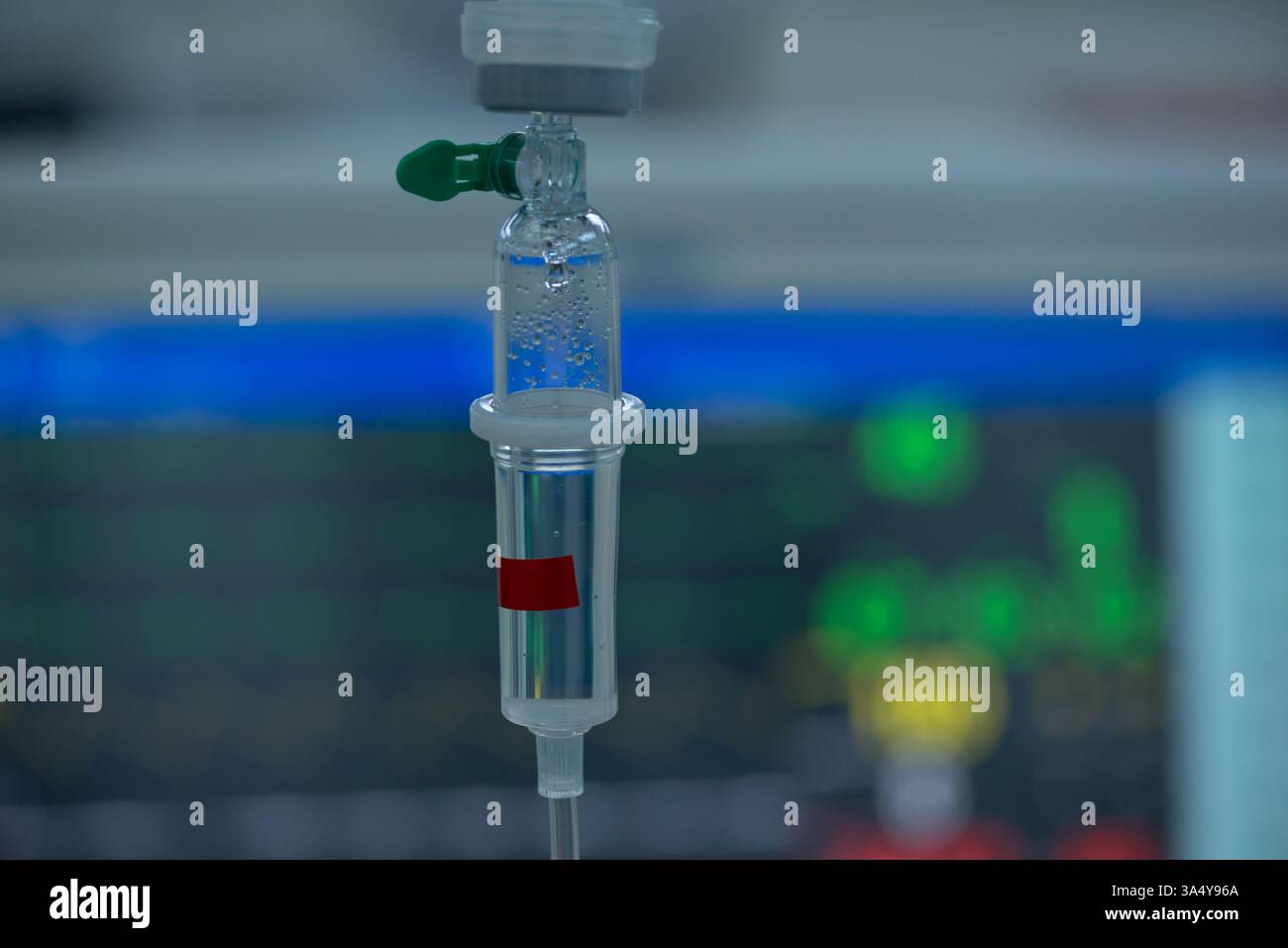 Immagine ravvicinata di una camera di gocciolamento endovenosa (IV) con bolle d'aria, posizionata su uno sfondo sfocato di un monitor medico che visualizza i segni vitali in una Foto Stock