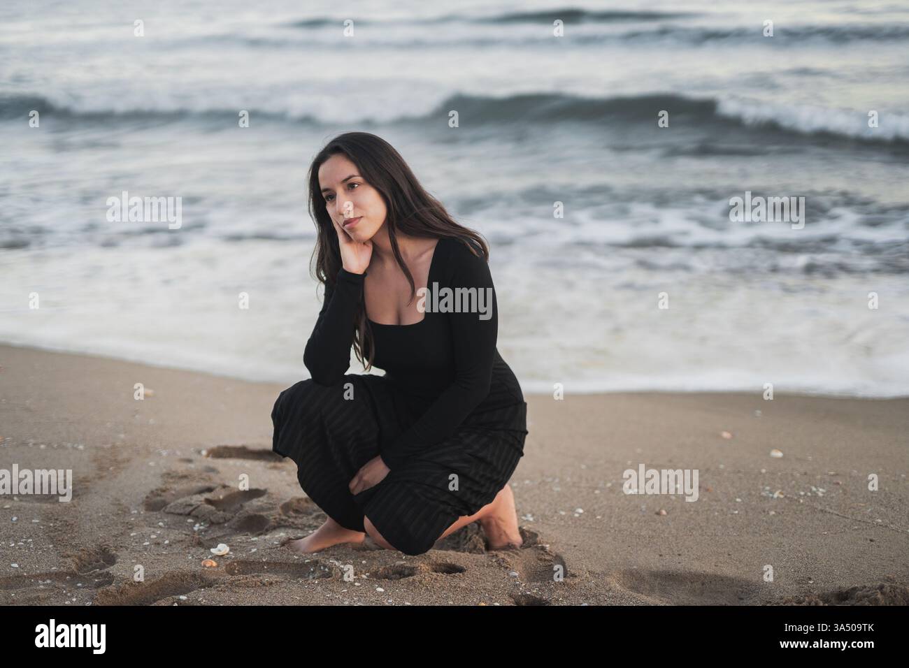Donna mista con lunghi capelli scuri che indossa un maxi abito nero si accosta su una spiaggia sabbiosa durante le vacanze. Guarda lontano con un'espressione calma e contemplativa, offrendo un'atmosfera serena di stile di vita di viaggio. Foto Stock