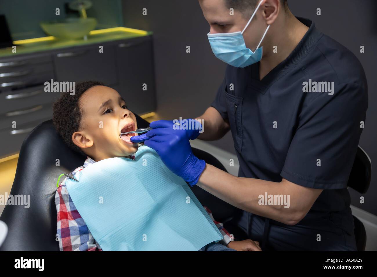 Scena al chiuso di un ragazzo dalla pelle scura al dentista, che sembra spaventato mentre il medico controlla i suoi denti. Questa immagine odontoiatrica pediatrica funziona bene per l'assistenza sanitaria, le cliniche odontoiatriche e le comunicazioni mediche familiari. Foto Stock