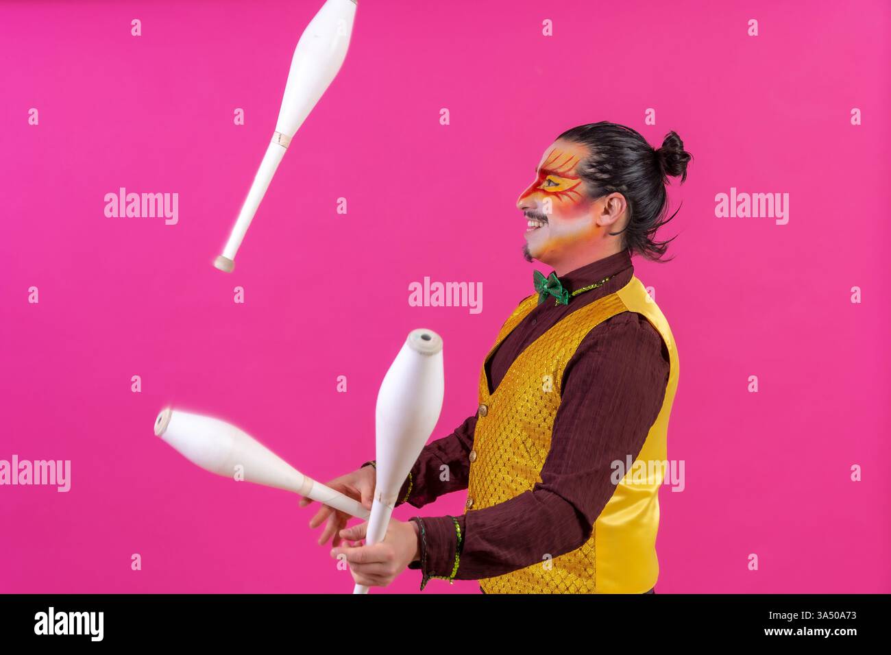 Clown con trucco facciale bianco sorride mentre si destreggia con maces sullo sfondo rosa di uno studio. Un'audace immagine a tema circense perfetta per l'intrattenimento, le prestazioni e i progetti spaziali. Foto Stock