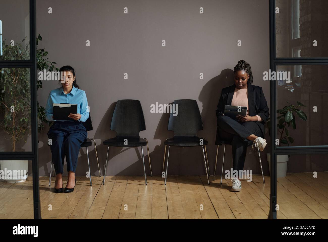 Due donne siedono in un corridoio compilando un modulo prima di un colloquio di lavoro durante il periodo della pandemia. L'immagine trasmette la messa a fuoco e la determinazione in un ambiente diversificato e professionale. Adatto a temi di reclutamento, risorse umane e occupazione. Foto Stock