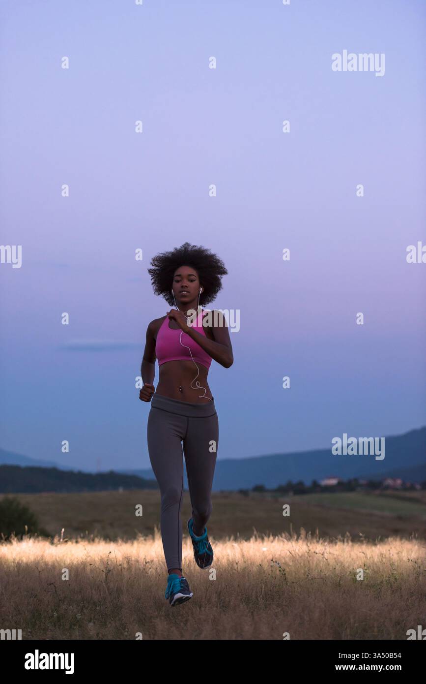 Giovane runner nera con cuffie che fa jogging all'aperto di notte attraverso un prato, catturando energia e vitalità. Ideale per campagne di fitness, benessere e attività all'aperto. Foto Stock