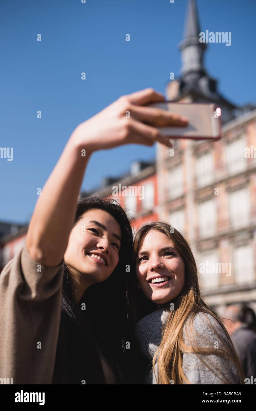 Gli amici cinesi ed europei fanno un allegro selfie a Plaza Mayor, Madrid. La scena all'aperto riflette l'amicizia interculturale e le visite turistiche della città in Spagna. Ideale per viaggi, stile di vita e momenti culturali. Foto Stock
