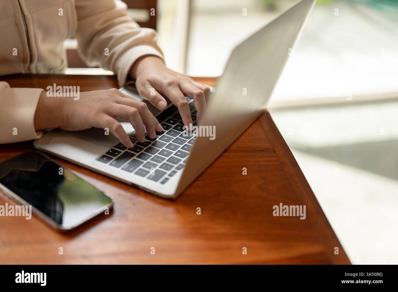 Primo piano di una giovane donna asiatica che usa un computer portatile in un tavolino di legno. Questa scena di lavoro a distanza si adatta ai concetti di freelance, coworking e nomadi digitali. Ideale per contenuti sul lavoro online, lo studio e la moderna cultura dei caffè. Foto Stock