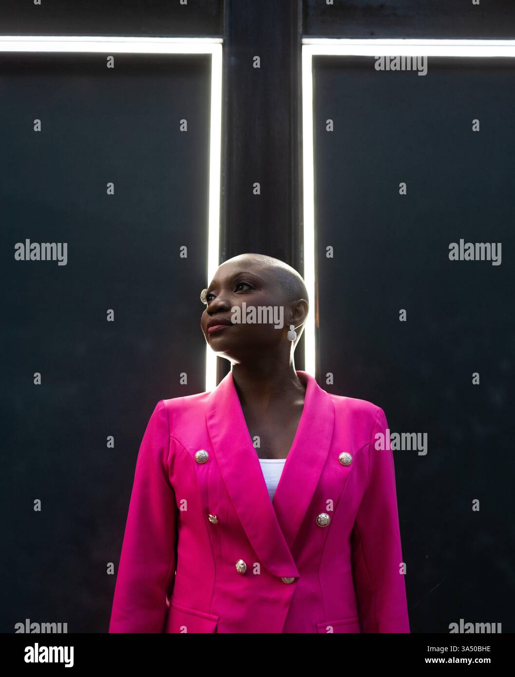 Ritratto ad angolo basso di una donna afroamericana sicura di sé con la testa rasata, indossa un abito rosa e orecchini. Si trova di fronte a cornici piene di luce, proiettando forza e individualità. Adatto per campagne di moda, empowerment e ritratti contemporanei. Foto Stock
