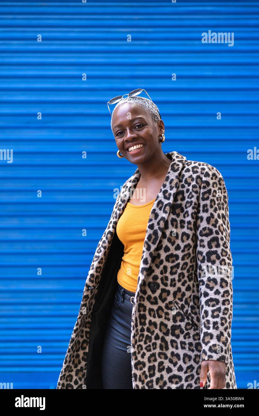 Donna nera sorridente con i capelli corti si staglia contro un muro blu in un ambiente urbano diurno. Questo luminoso ritratto urbano si adatta allo stile di vita, alla moda e alle campagne di benessere. Foto Stock