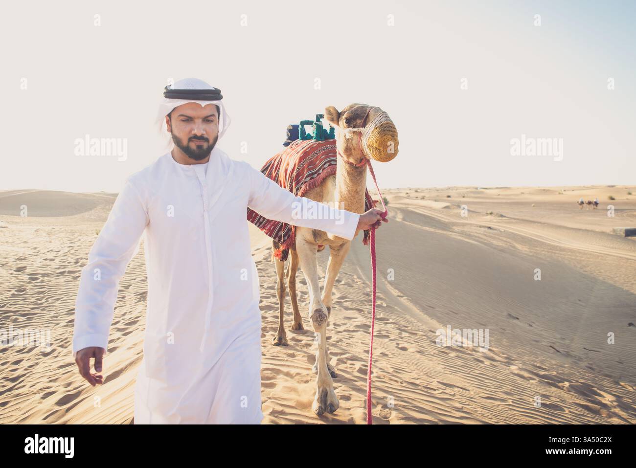 Uomo con abiti tradizionali che conduce un cammello attraverso il deserto di Dubai. Questa immagine dello stile di vita del viaggio mette in evidenza paesaggi desertici, abbigliamento regionale e atmosfera da safari nel Golfo. Adatto per viaggi, cultura e temi del patrimonio mediorientale. Foto Stock