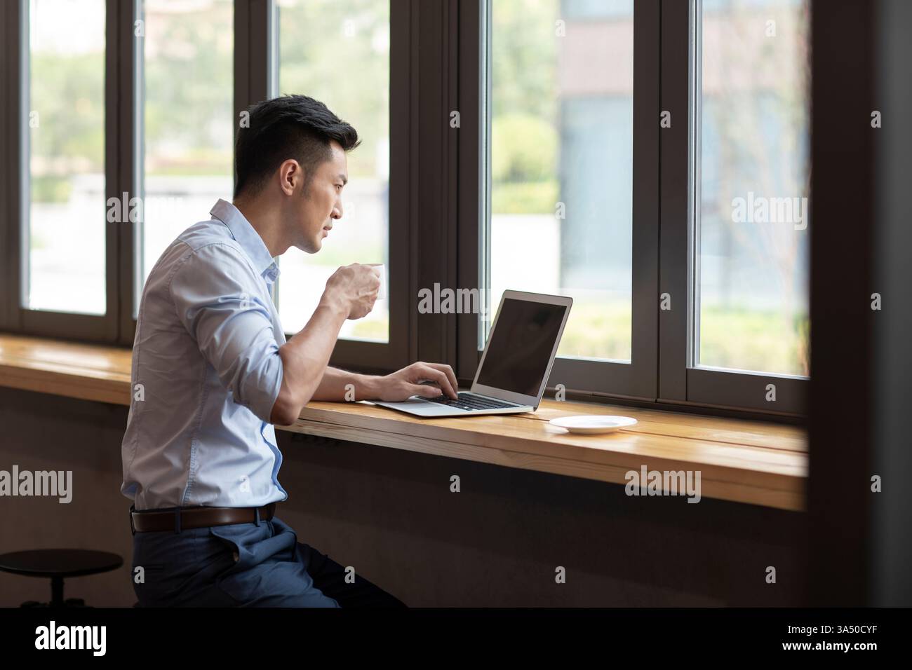 L'imprenditore cinese maschile in un bar utilizza un computer portatile e un caffè, catturati in un momento di lavoro informale. Ideale per storie di lavoro remoto, business online e moderni spazi di lavoro urbani. Foto Stock
