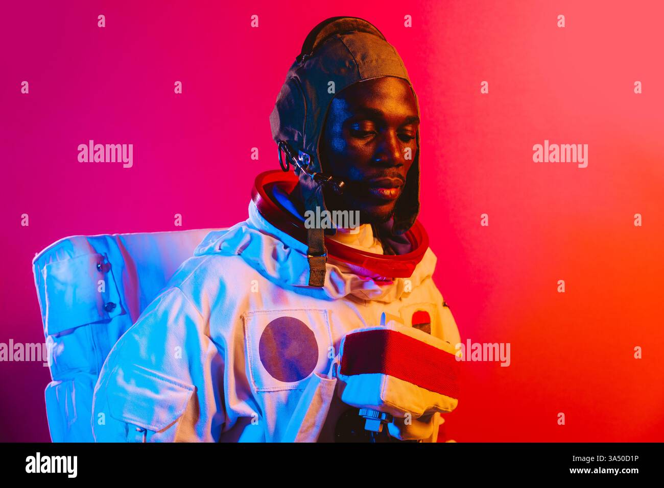 Uomo afro-americano in tuta spaziale che guarda verso il basso su uno sfondo di studio dai colori vivaci. Questo ritratto a tema scientifico e spaziale funziona bene per la tecnologia, l'educazione scientifica e le campagne di ispirazione. Ideale per l'esplorazione dello spazio e la narrazione STEM. Foto Stock