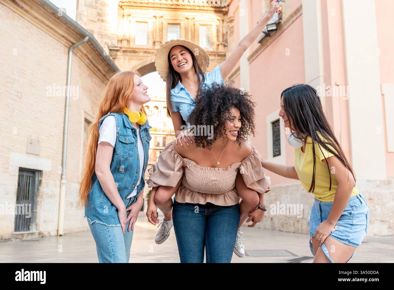 Gruppo di donne multirazziali in una strada di Valencia sorridente, con un amico che fa un giro in piggyback. Ideale per viaggi, amicizia, stile di vita urbano e campagne estive. Foto Stock