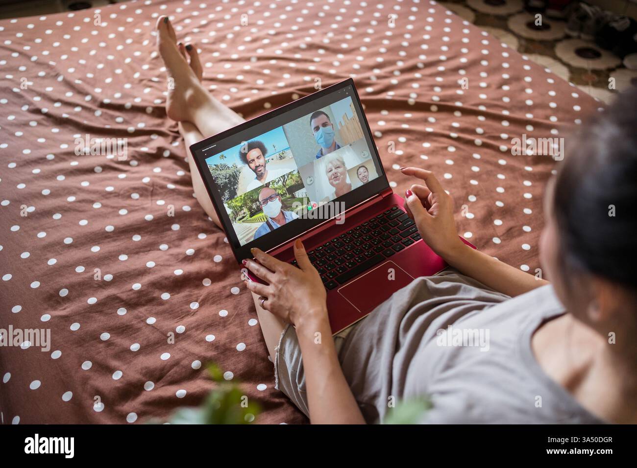 La donna asiatica si siede su un letto a casa e videochatta con amici di diverse origini. Questa scena mette in evidenza la connessione remota e la comunicazione online con un gruppo multirazziale. Ideale per i contenuti che riguardano la connessione durante il nuovo normale distanziamento sociale e stili di vita digitali. Foto Stock