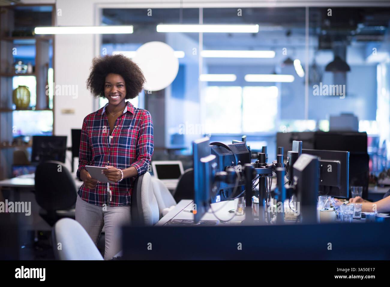 Donna d'affari nera sorridente con Afro che tiene in mano un tablet in un moderno ambiente d'ufficio. Immagini professionali per lo stile di vita aziendale, ideali per campagne tecnologiche, di leadership e sul luogo di lavoro. Adatto per progetti a tema business, imprenditorialità e successo. Foto Stock