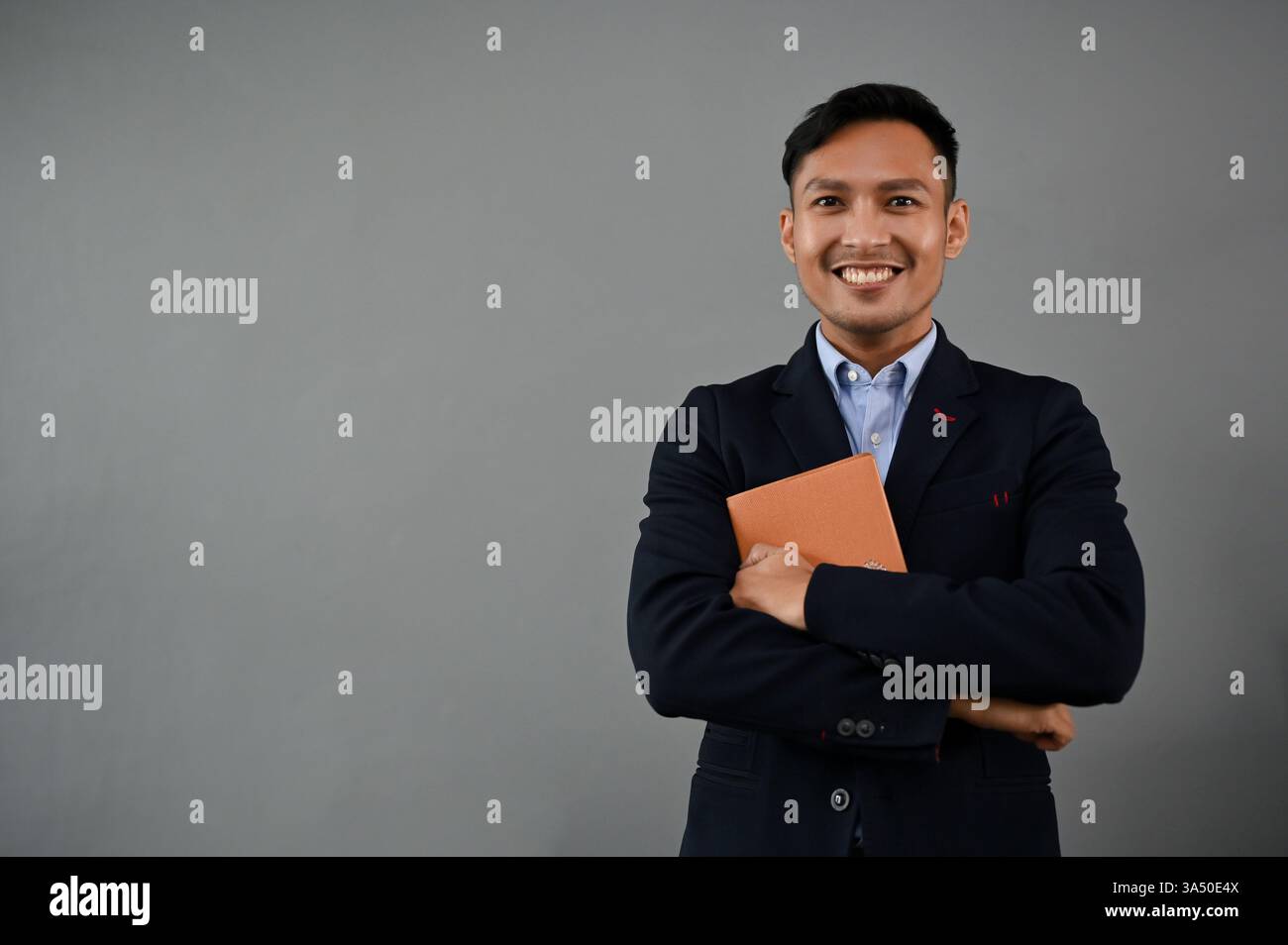 L'uomo d'affari asiatico, sorridente e millenario in una tuta formale, regge un libro sullo sfondo di uno studio grigio neutro. Questo ritratto professionale si adatta alle comunicazioni aziendali, alla leadership e al branding aziendale. Ideale per campagne con professionisti asiatici o dirigenti thailandesi. Foto Stock