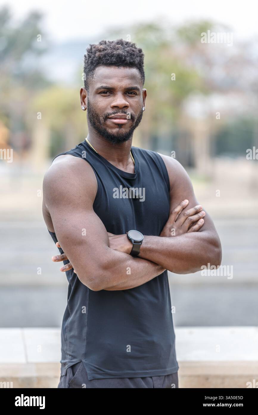 Un uomo afroamericano serio nell'abbigliamento sportivo si erge con le braccia incrociate in un parco illuminato dal sole. L'ambiente all'aperto enfatizza il fitness, il benessere e uno stile di vita attivo. Ideale per campagne di formazione, motivazione e vita sana. Foto Stock