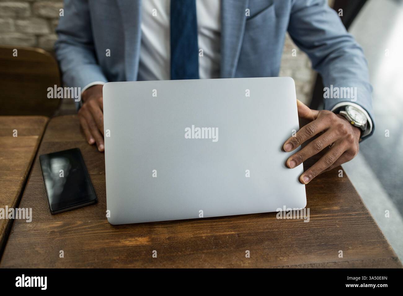 Un uomo d'affari di colore in tuta inizia a lavorare su un computer portatile presso una scrivania di legno in un ufficio moderno. Un momento professionale che enfatizza la tecnologia, il business e la vita lavorativa aziendale. Ideale per le comunicazioni aziendali, l'imprenditorialità e i temi del lavoro online. Foto Stock