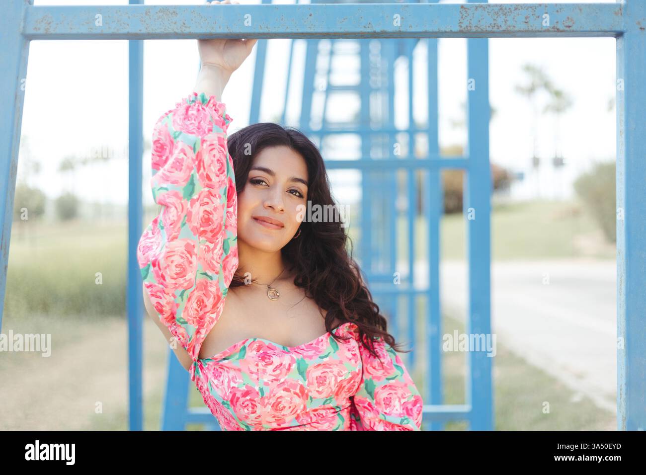 Una donna ispanica sorridente che indossa un top a spalla rosa con motivi floreali si staglia all'aperto con una montatura metallica in una giornata di sole. Il luminoso ambiente esterno e la sua espressione allegra trasmettono un vivace umore estivo ideale per la moda, lo stile di vita o le campagne di parchi. Ideale per promuovere lo stile casual, la positività e le immagini all'aperto. Foto Stock