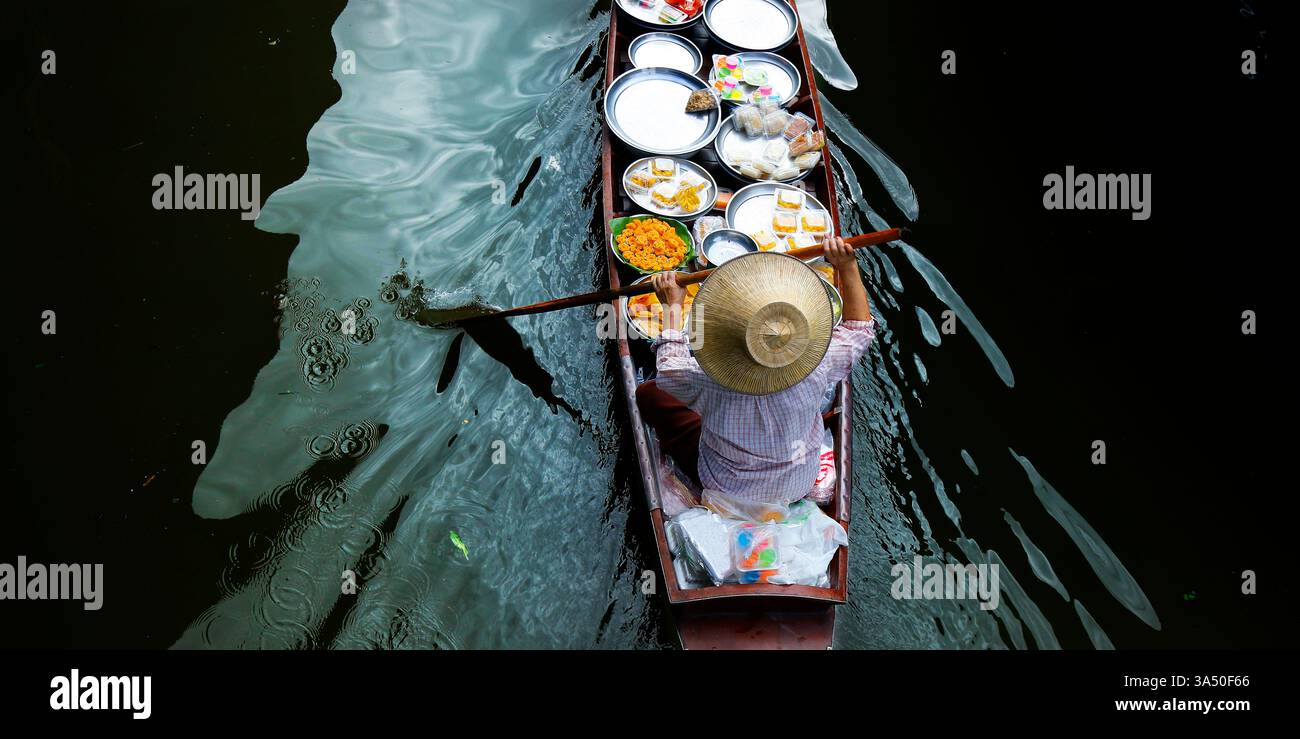 Scena colorata al mercato galleggiante di Damnoen Saduak con una donna in cappello a bordo di una barca carica di prodotti freschi. L'immagine cattura la tradizionale vita del mercato tailandese sul canale con barche, frutta e verdura in vendita. Nessun volto visibile, il che lo rende versatile per le campagne di viaggio, cultura e lifestyle. Foto Stock
