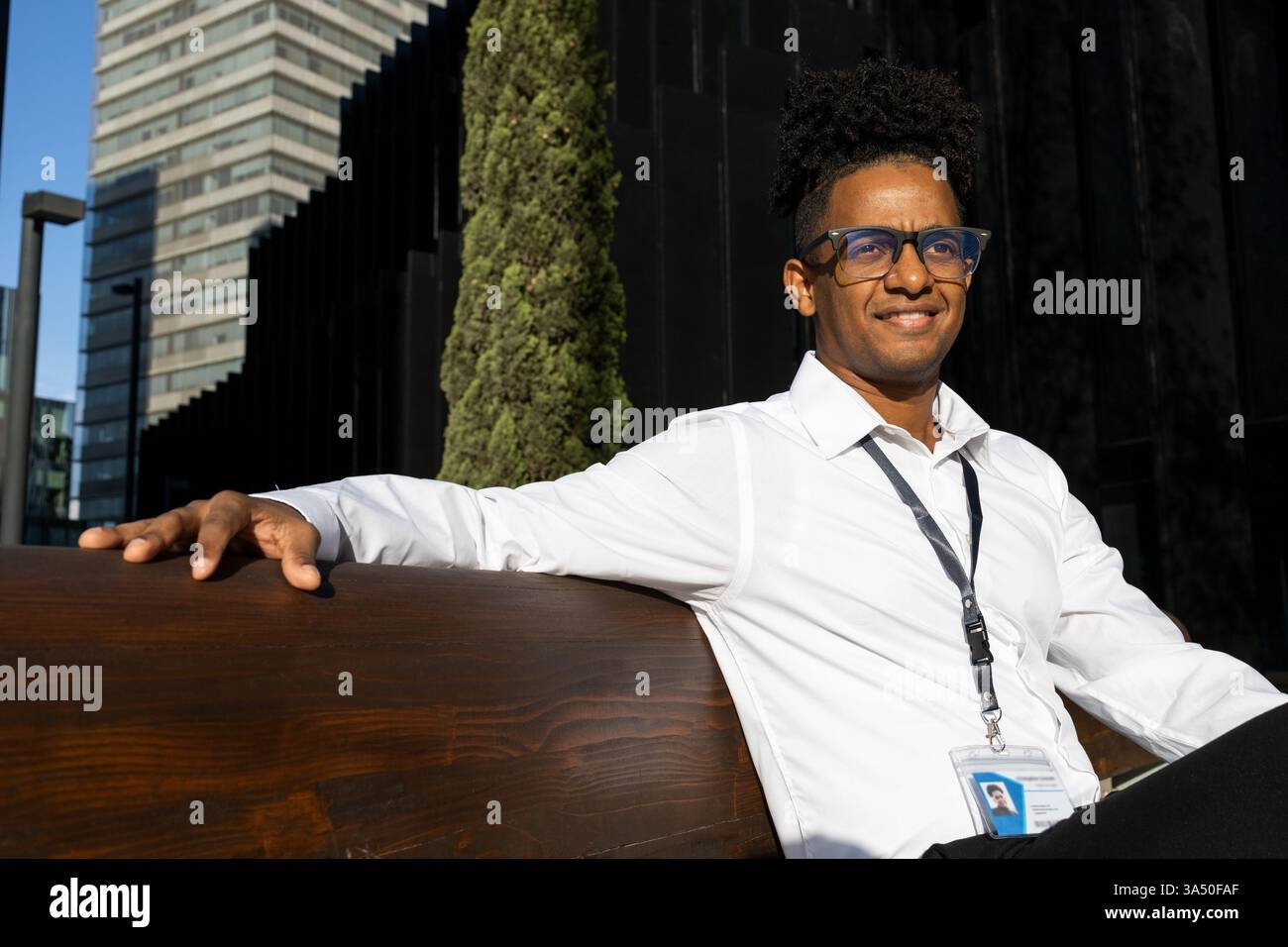 Uomo afroamericano positivo in una camicia bianca seduta su una sedia di cemento su una strada soleggiata. Un ritratto urbano all'aperto che comunica sicurezza e stile. Foto Stock