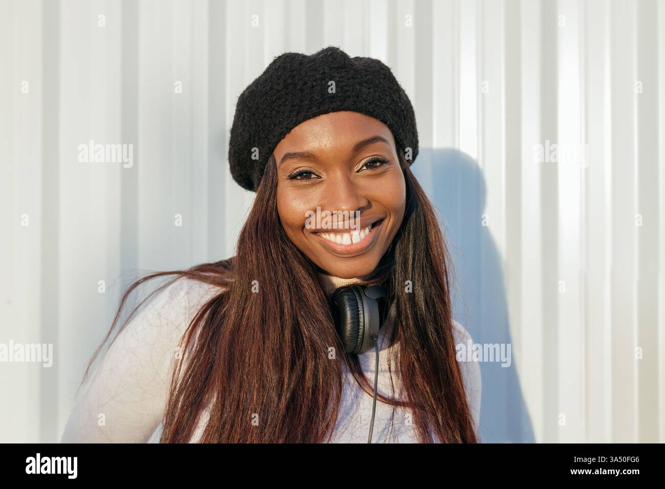 Allegra donna nera che indossa le cuffie poggia all'aperto contro una parete a coste illuminata dal sole. Il ritratto informale cattura la positività e un'atmosfera accessibile con luce naturale e un sorriso caldo. Perfetto per lo stile di vita, la musica o le campagne di moda urbana. Foto Stock
