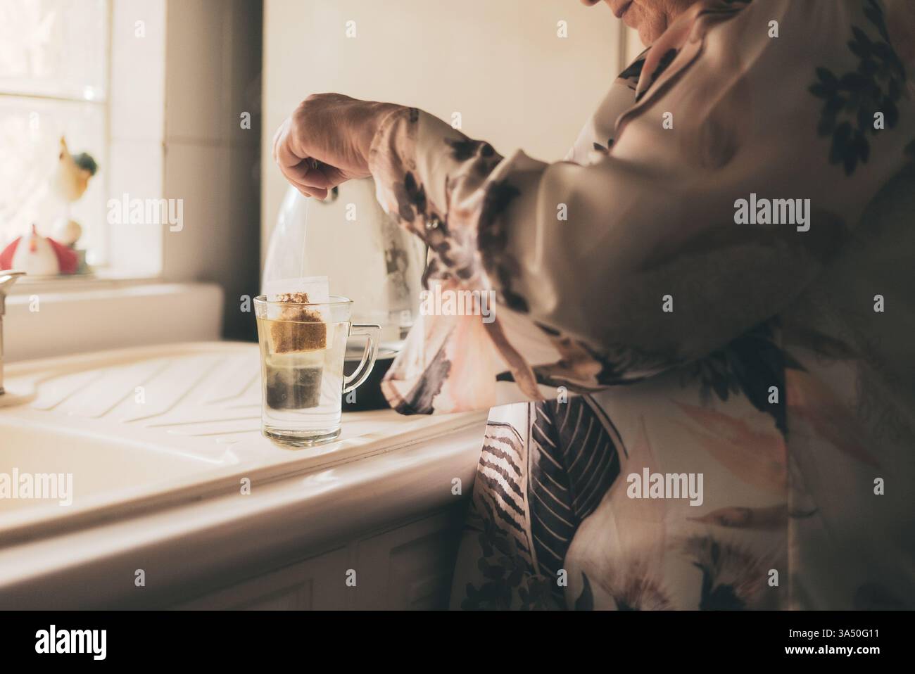 Donna anziana ispanica che prepara il tè in una tazza di vetro in cucina a casa. Un'immagine calda e invitante, perfetta per l'invecchiamento, la routine domestica e i momenti di comfort. Utile per contenuti senior o di ospitalità. Foto Stock