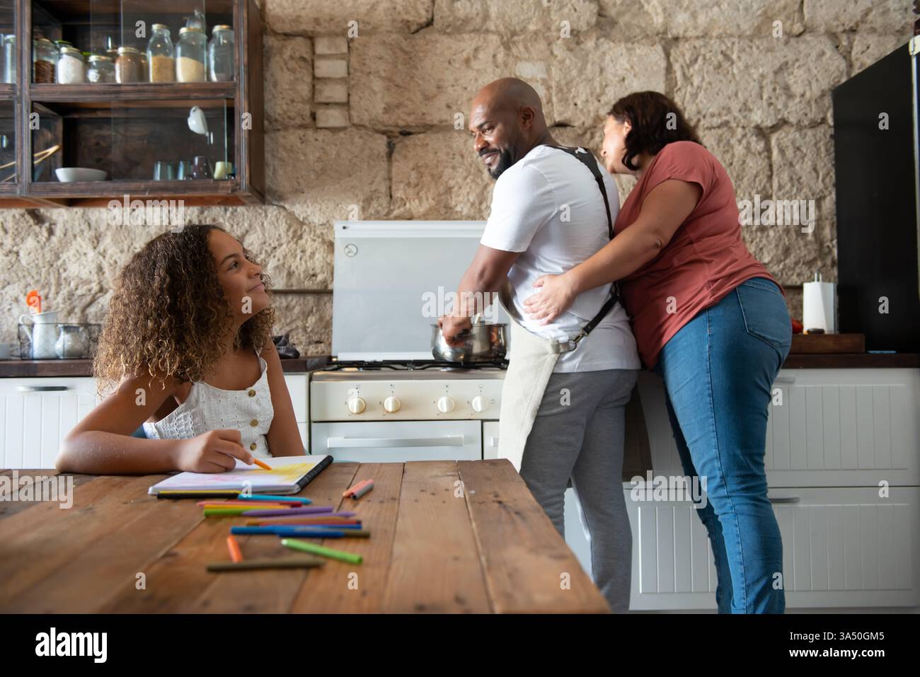Il padre Nero sorridente cucina mentre sua moglie lo abbraccia da dietro e guarda verso la figlia al tavolo. Pastelli colorati e quaderni aggiungono un tocco familiare in cucina. Perfetto per le campagne di vita familiare, genitorialità e stile di vita domestico. Foto Stock