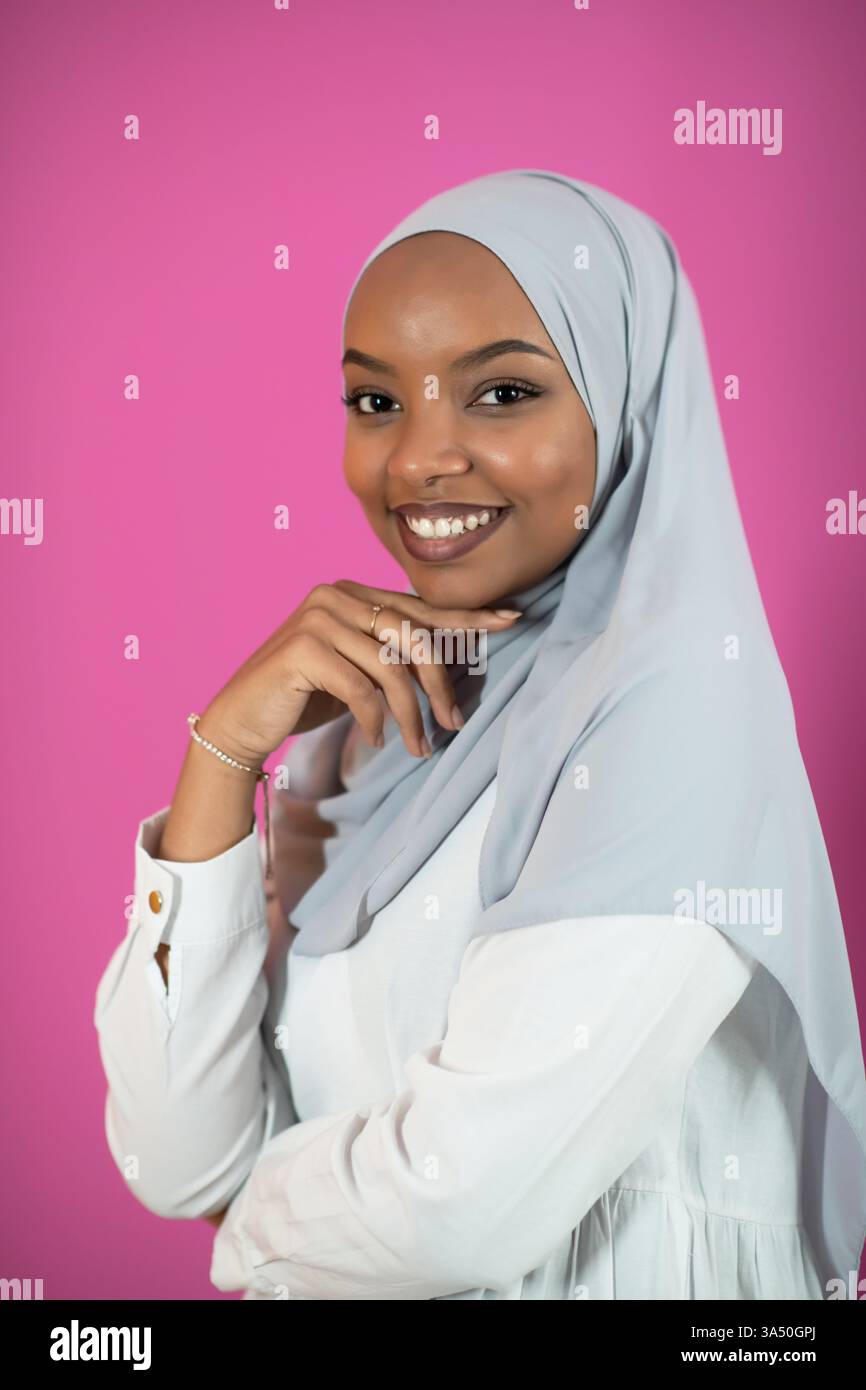 Giovane donna afro che indossa abiti islamici tradizionali su uno sfondo di plastica rosa. Una rappresentazione vibrante della moda, della cultura e della diversità islamiche, ideale per campagne di stile di vita e bellezza. Lo sfondo rosa tenue migliora il calore e l'eleganza. Foto Stock