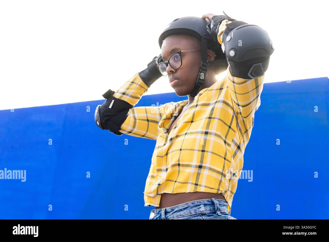 Vista dall'angolo basso di una premurosa donna afroamericana che regola un casco contro una parete blu e un cielo limpido. Ideale per pattinaggio in linea, equipaggiamento sportivo di sicurezza e attività ricreative all'aperto. Foto Stock