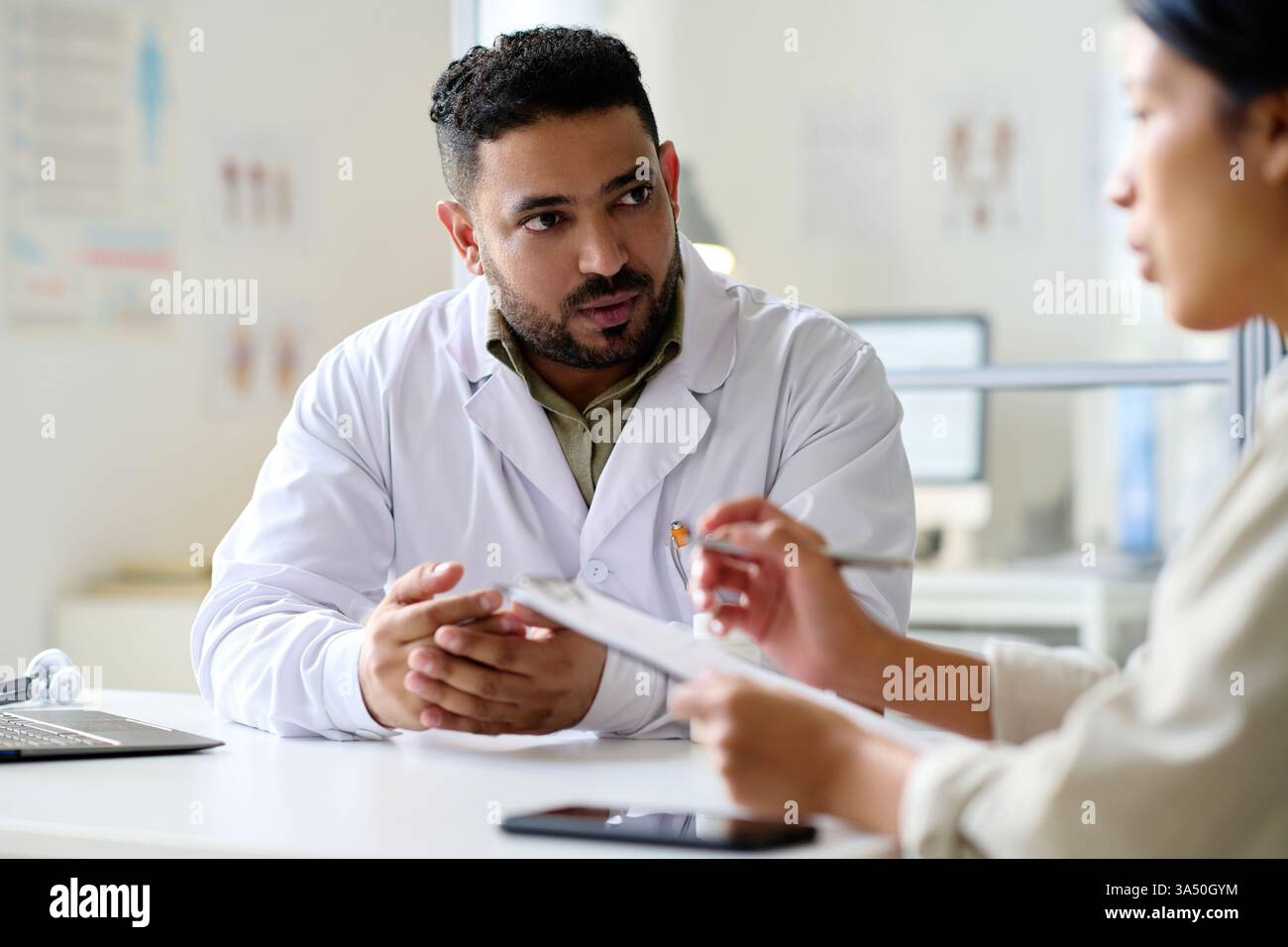 Scena medica che mostra un medico discutere di trattamento con un paziente che sta firmando un modulo di consenso in un ambiente ospedaliero. L'interazione professionale si adatta ai temi della comunicazione sanitaria, clinica e paziente. Include una scrivania e un computer portatile per enfatizzare le moderne cure mediche. Foto Stock