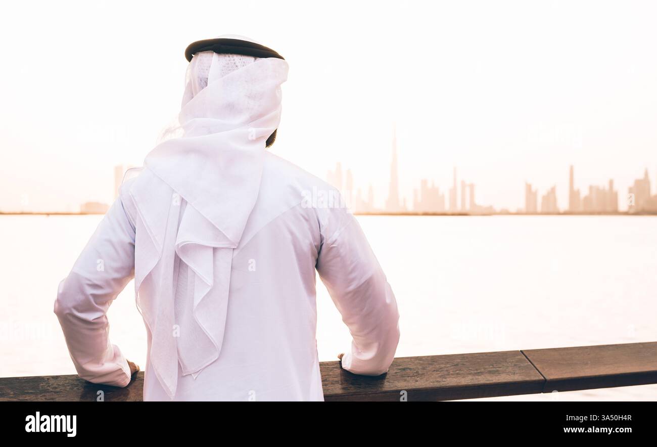 Uomo mediorientale che indossa kandura appoggiato sulla recinzione di fronte al mare al tramonto Foto Stock