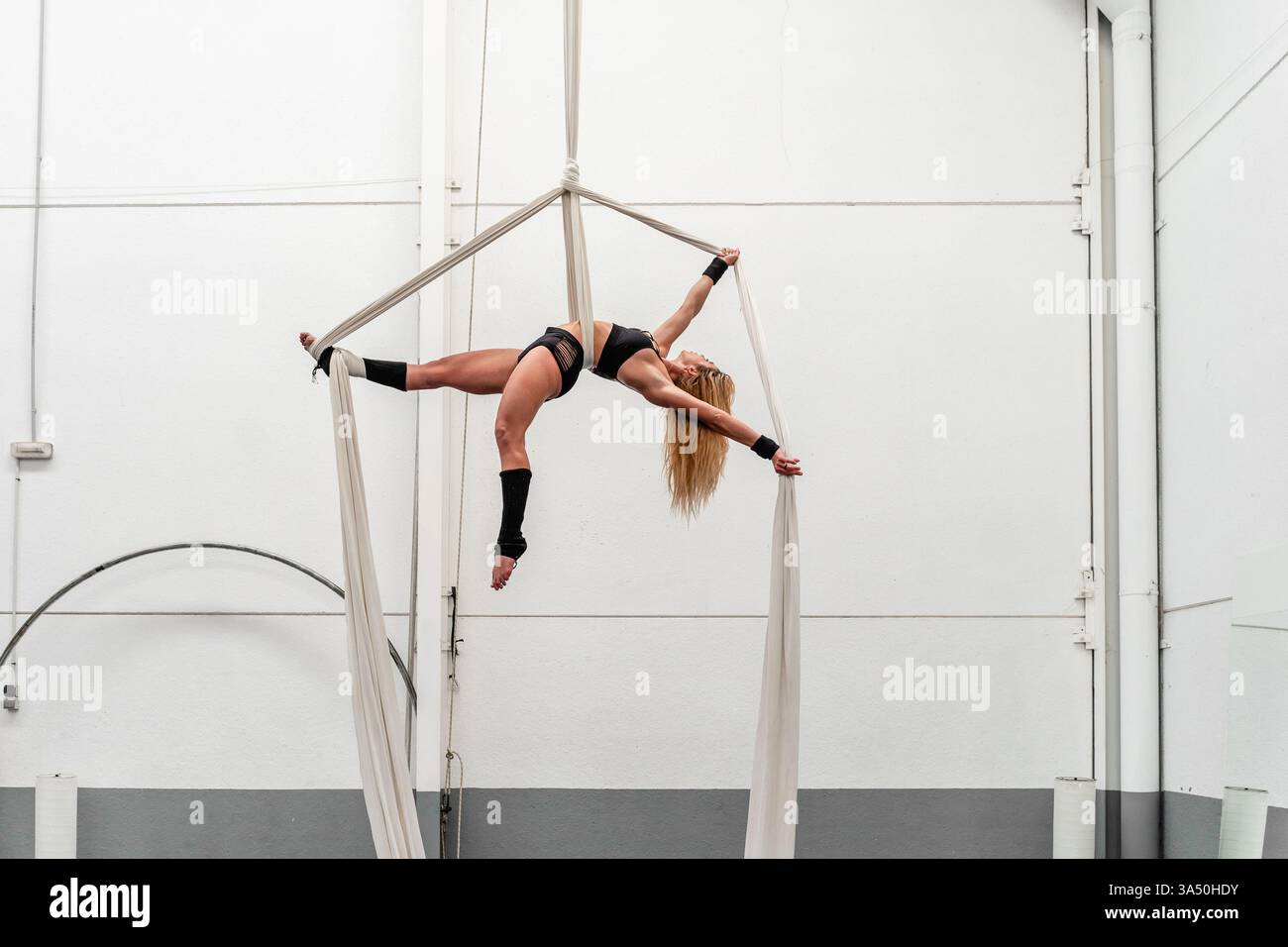 Donna ispanica che pratica sete aeree in un centro fitness, posa a mezz'aria che mostra flessibilità e forza. L'ambiente della palestra mette in evidenza acrobazie, danza e allenamento fitness. Eccellente per campagne di benessere, sport e stile di vita attivo. Foto Stock