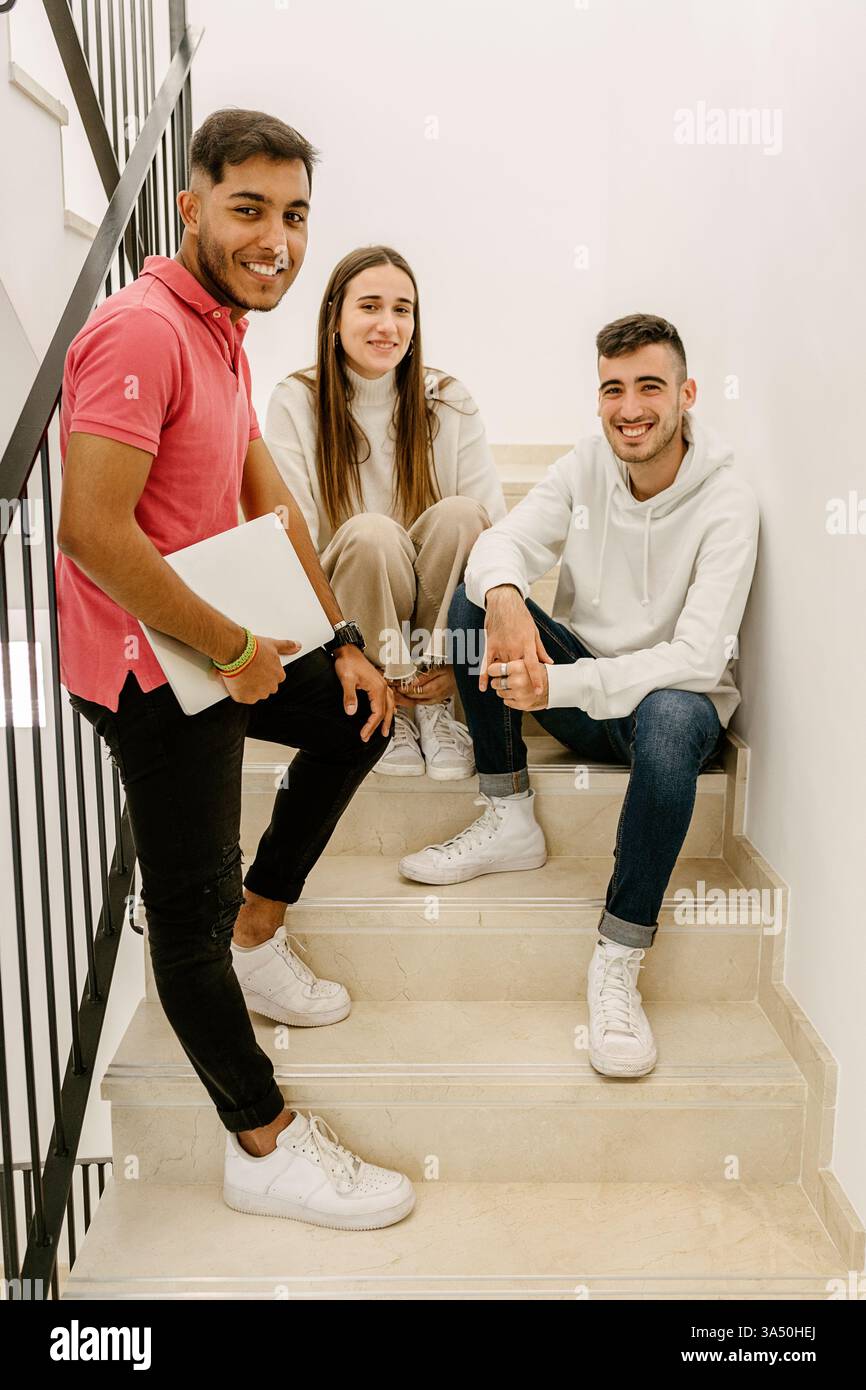 Un uomo ispanico con un portatile siede su una scala con gli amici, appoggiato alla ringhiera. Un'immagine di stile di vita studentesco facile e adatta a temi educativi, culturali giovanili e di collaborazione. Foto Stock