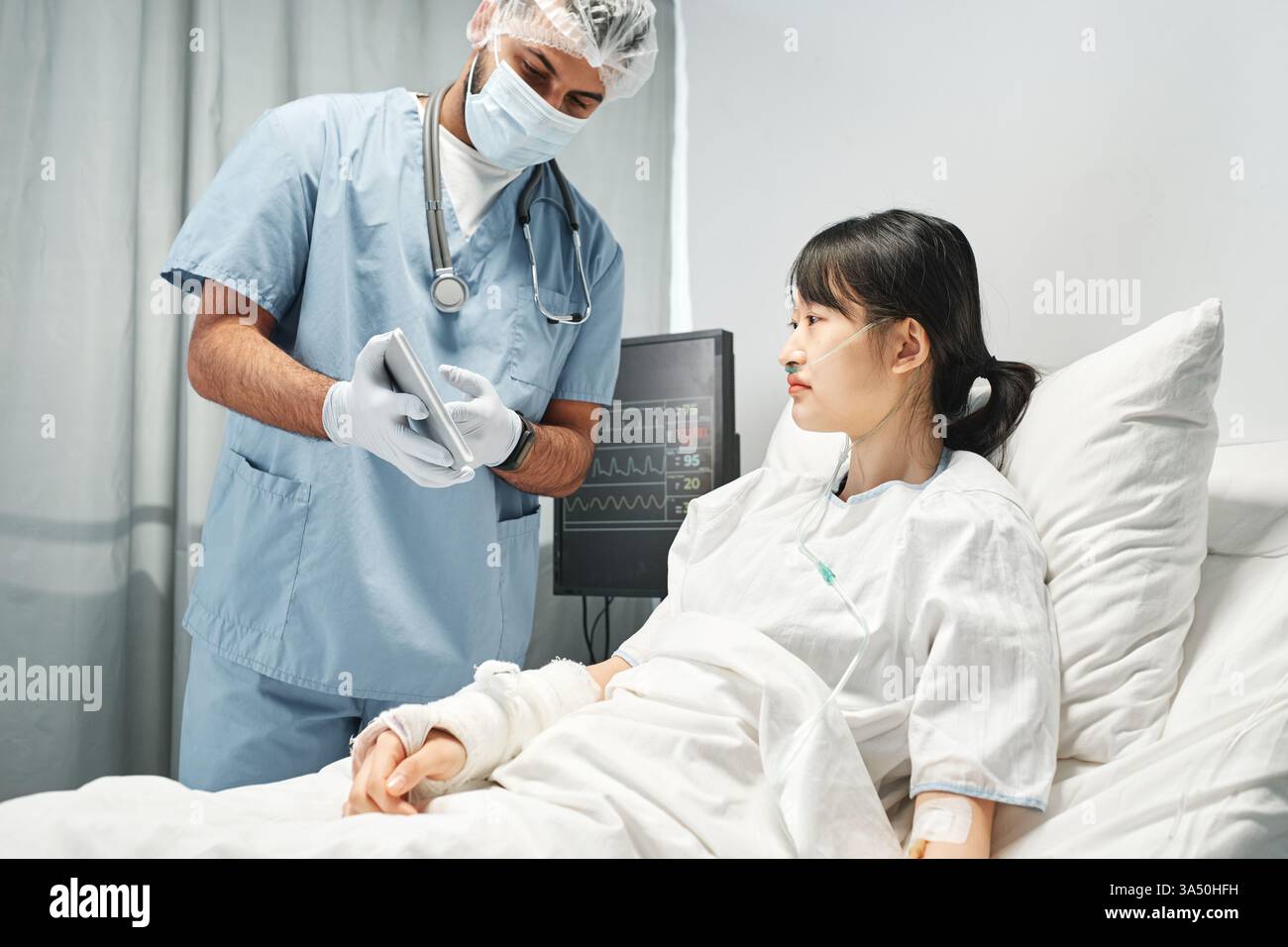 Operatore sanitario che indossa una maschera dimostra qualcosa su un tablet digitale per un giovane paziente asiatico. Questo momento cattura l'interazione tra medico e paziente in un ambiente clinico e mette in evidenza l'assistenza medica e la comunicazione. Ideale per i contenuti di educazione sanitaria, ospedaliera o medica. Foto Stock