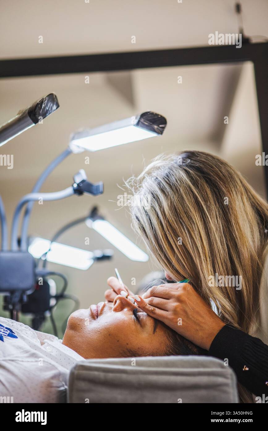Estetista ispanico che esegue precise estensioni delle ciglia per un cliente in un salone moderno. Il primo piano cattura la cosmetologia professionale e la cura prestata durante un trattamento delle ciglia. Foto Stock