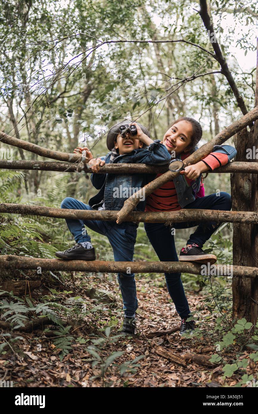 Ragazzo e ragazza ispanica con binocolo appoggiato contro un ramo di albero in una foresta. Un affascinante scenario di esplorazione all'aria aperta per attività familiari ed educazione alla natura. Ideale per l'apprendimento dei bambini, l'ecologia e la grafica di avventura nei boschi. Foto Stock