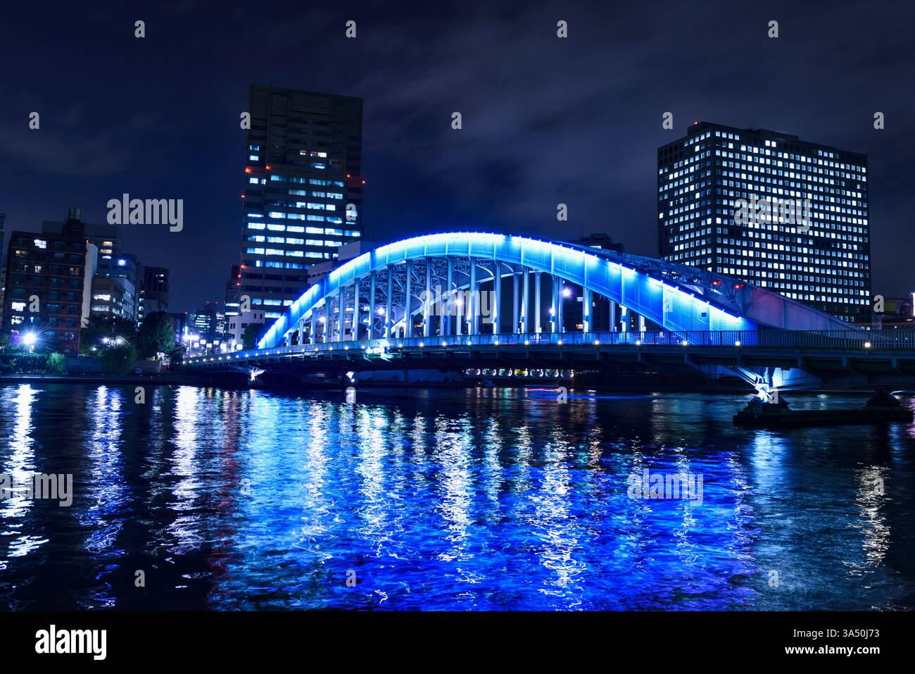 Vista notturna di Tokyo con il fiume Sumida e il Ponte Eitai illuminati contro uno skyline di alti edifici. Ideale per viaggi, fotografie notturne in città e caratteristiche architettoniche. Ottimo per la copertura della vita cittadina di Tokyo e dei paesaggi urbani con riflessi sull'acqua. Foto Stock