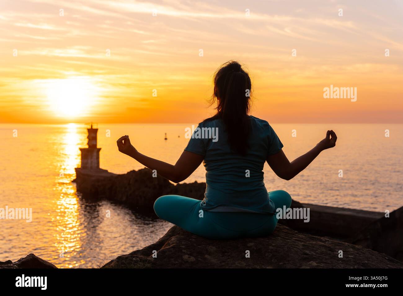 Silhouette di una donna che meditava su una roccia al tramonto accanto a un faro sul mare. Un'immagine di benessere tranquillo che trasmette equilibrio e uno stile di vita naturista. Foto Stock