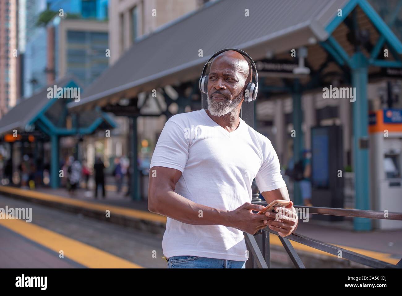 Un afroamericano con indosso una camicia scura e le cuffie aspetta in una stazione ferroviaria del New Jersey. Ascolta la musica sul suo telefono cellulare, catturando un momento di viaggio in città all'aperto in una giornata di sole. Questa immagine è ideale per il trasporto, i viaggi e il marketing lifestyle. Foto Stock