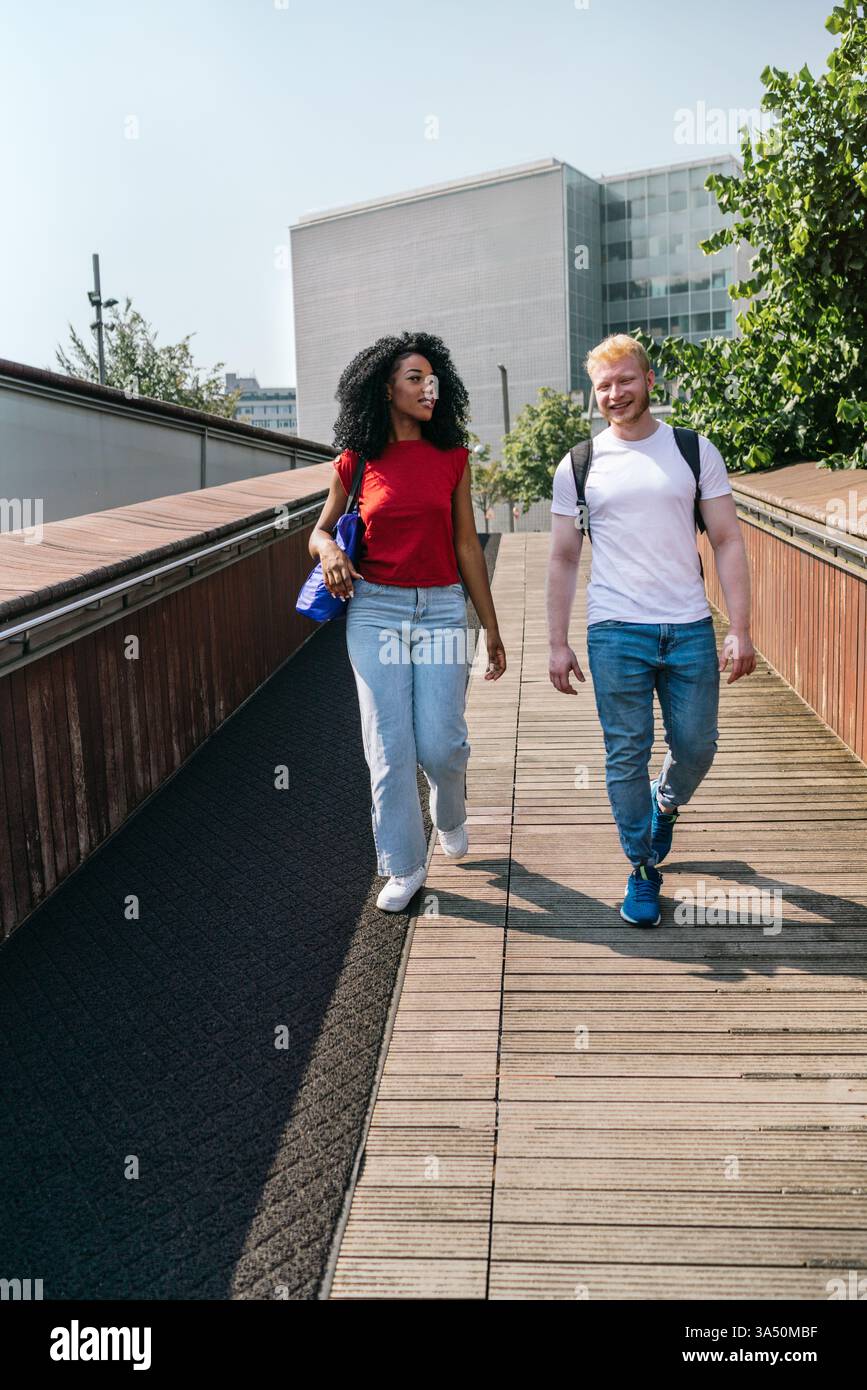 Due giovani adulti in una coppia multirazziale passeggiano su una passerella di legno in città in una giornata di sole. Chiacchierano e sorridono, trasmettendo un rapporto caldo e uno stile di vita urbano di incontri. Un'immagine versatile per lo stile di vita, il romanticismo e le campagne di vita in città. Foto Stock