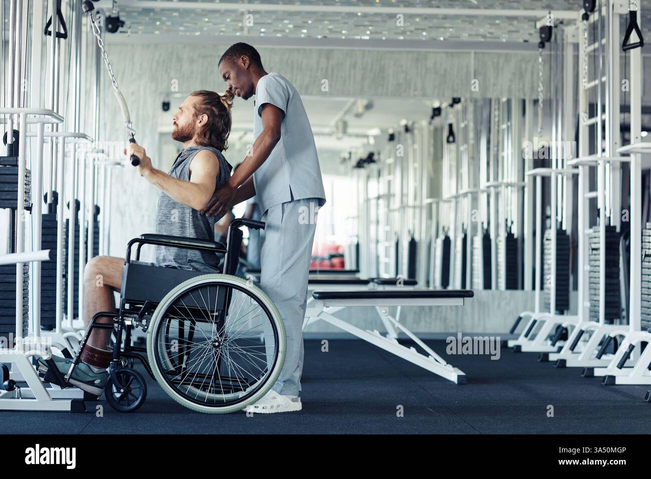 Paziente giovane con disabilità seduto su una sedia a rotelle, sostenuto da un medico durante la riabilitazione. Questo ambiente sanitario mette in evidenza l'inclusione della disabilità, la fisioterapia e il recupero, adatto per campagne mediche o di benessere. Foto Stock