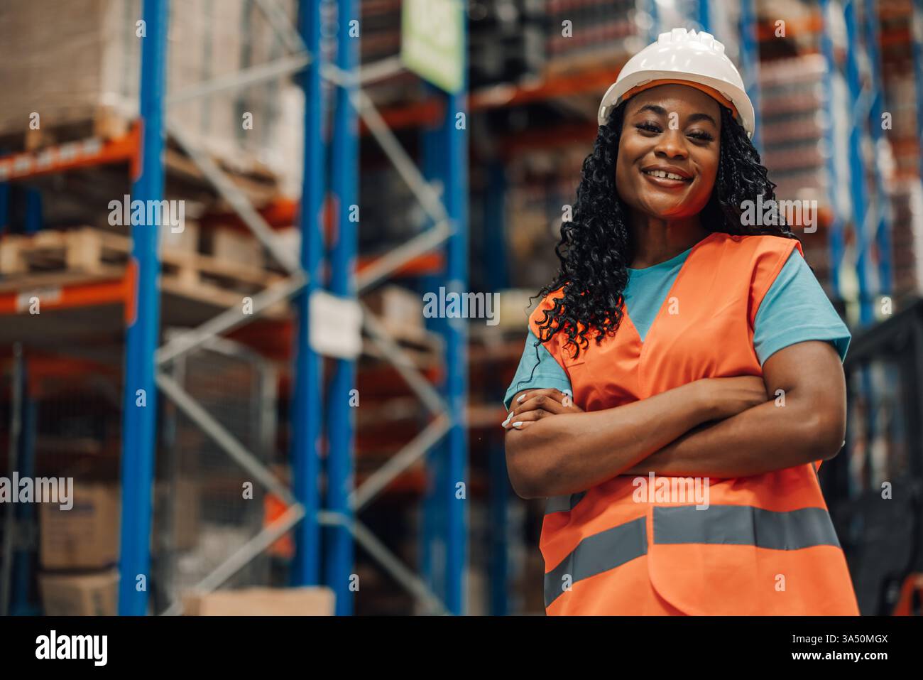 Con le braccia incrociate, una donna afro-americana sorridente con gli attrezzi di sicurezza del magazzino trasmette fiducia e orgoglio. Questo ritratto industriale mostra le donne che operano nella logistica e negli ambienti di produzione moderni. Foto Stock