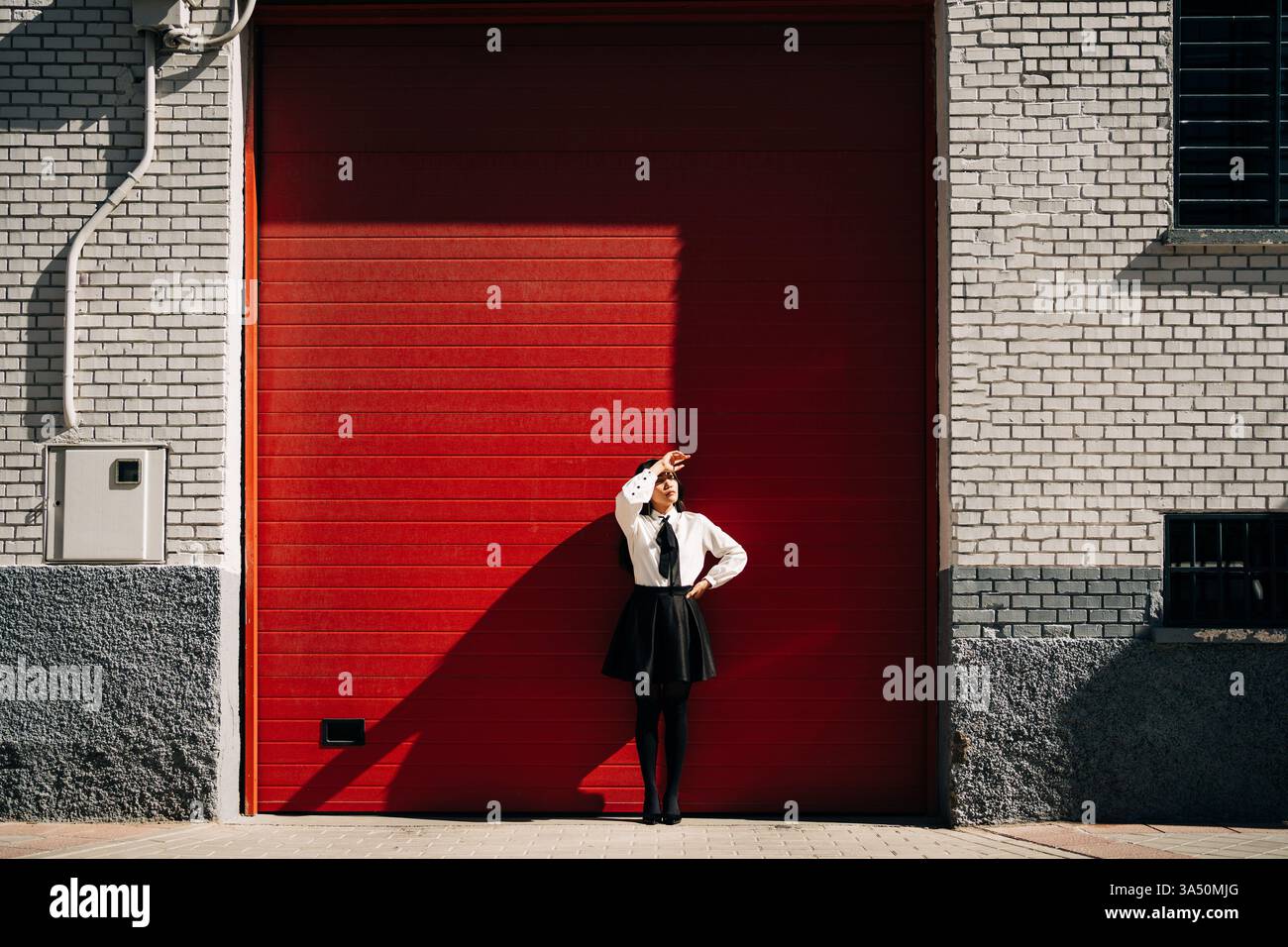 Donna asiatica con una mano sulla vita che protegge il viso dal sole, in piedi all'aperto in un ambiente urbano luminoso. La luce e l'ombra audaci creano un look elegante e contemporaneo. Adatto a contesti di moda, estate e vita all'aperto. Foto Stock