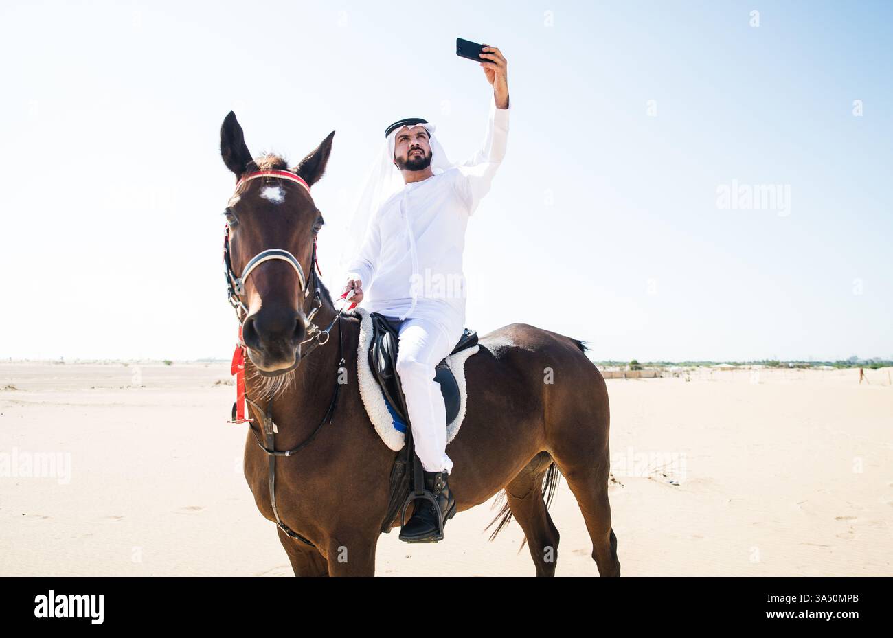 Un giovane che indossa una kandura cavalca su un cavallo attraverso il deserto, catturando un momento di stile di vita degli Emirati. La scena mescola l'abito tradizionale con l'ampio paesaggio desertico, ideale per viaggi, cultura e caratteristiche di lusso. Adatto per articoli sul Medio Oriente, attività equestri o patrimonio regionale. Foto Stock