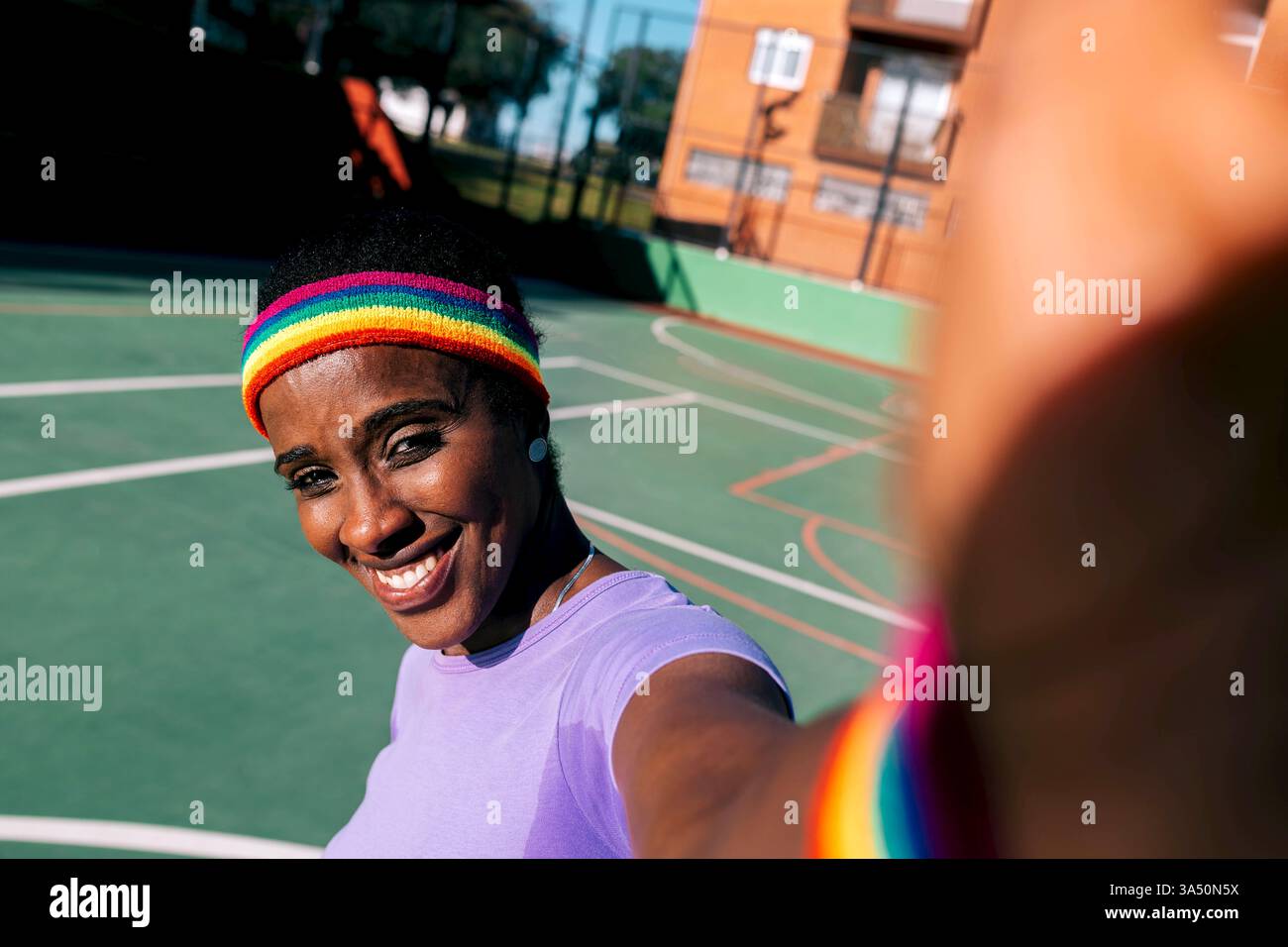 Dall'alto, allegra donna afroamericana in abiti casual sorride per un selfie su un campo da stadio. Ha uno smartphone in testa per un momento sociale ad alta energia. Ideale per lo sport, il tempo libero e le immagini dello stile di vita. Foto Stock